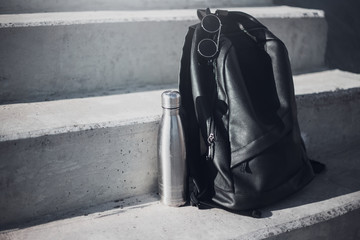 Reusable steel thermo water bottle, round sunglasses and black backpack on urban stairs.