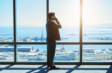 Fototapeta premium Mature executive at large windows on phone with harbour view