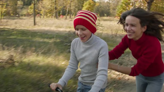 Young Caucasian woman teaches her son to ride a scooter in the autumn forest.