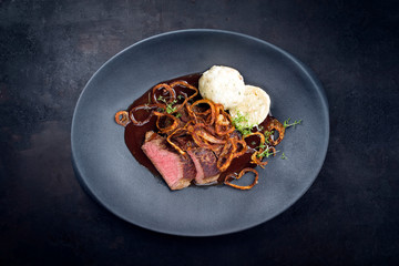 Traditioneller dry aged Rostbraten mit Röstzwiebel Kartoffel Knödel und Bratensauce serviert als Draufsicht auf einem Modern Design Teller