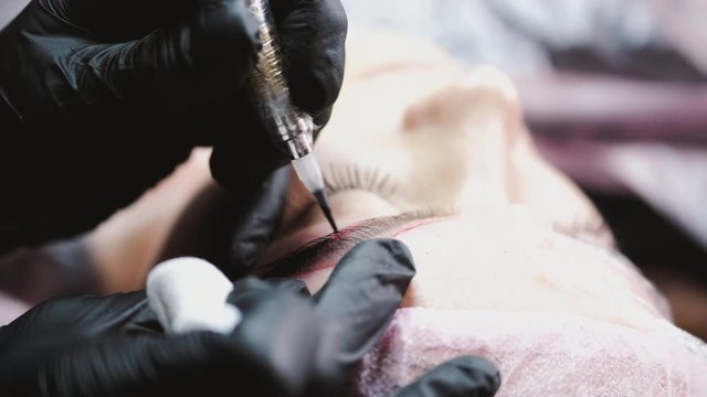 A beauty master in rubber gloves is dying the eyebrows of a client with the microblading technique.The professional is following the eyebrow mapping.