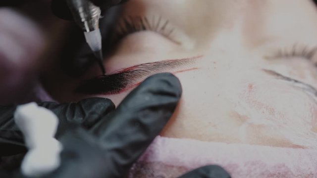 A beauty master in rubber gloves is dying the eyebrows of a client with the microblading technique.The professional is following the eyebrow mapping.