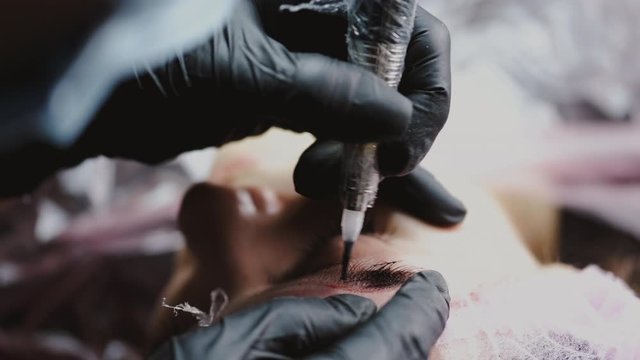 A professional brow artist is doing microblading on brows. The master is applying tint and a client is lying on a couch in a disposable hat.