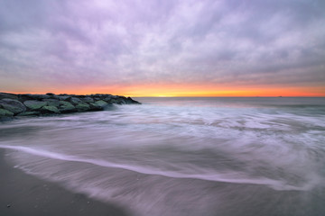 Dark ominous storm clouds and a sliver of golden light over a stone jetty on a beach, with swirling waves in motion. Long Beach, New York. 