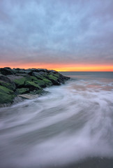 Dark ominous storm clouds and a sliver of golden light over a stone jetty on a beach, with swirling waves in motion. Long Beach, New York. 