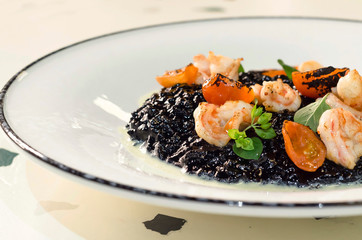rice with cuttlefish ink, octopus, mussels, squid, shrimp. prawns and grilled tomatoes on a large white plate in a restaurant. Italian black risotto with seafood. national dishes of italy.