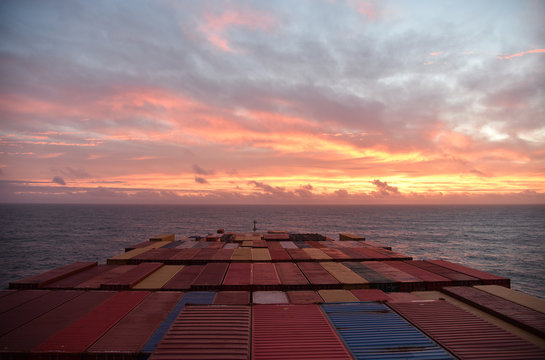 Container ship sailing through Atlantic Ocean toward beautiful sunrise. View on deck loaded with containers. 