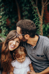 Obraz premium Little blonde girl sitting between mon and dad. Father kisses mother on the head. Daughter enjoying with her young parents in the garden. Love and family concept.