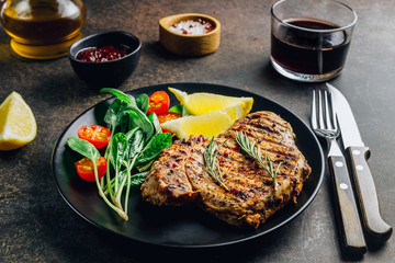 Roasted pork steak on black plate over dark background. With lemon, Cranberry Sauce and wine.