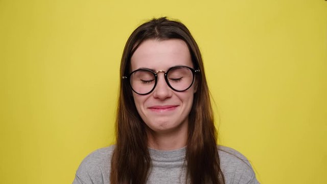 Cute young girl in eyeglasses makes funny face, crosses eyes and sticks out tongue, plays fool, doesnt want to be responsible and looking at camera, stands over yellow background. Positive emotions