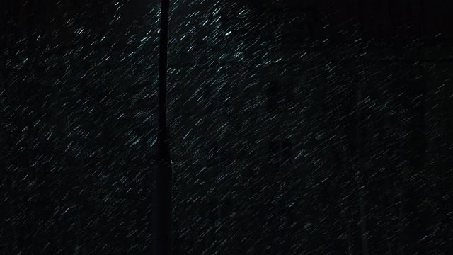 Night street with lanterns and luminous window in building opposite. Snowflakes falling in light of lantern. Luminous lettering, traffic lights, brake lights. Headlights of cars driving along street