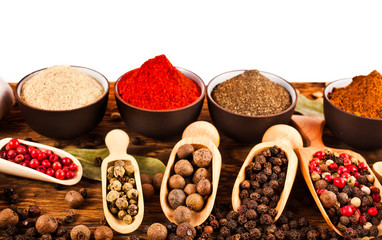 Different peppers in ceramic cups and peppercorns in wooden scoops on wooden background.