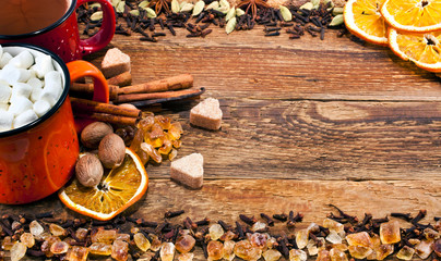 Coffee and fluffy white marshmallows in enamel cups with a variety of spices, dried slices of orange with cinnamon sticks, nutmeg and sugar hearts on wooden background. Text space. Greeting card.
