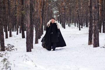 A warrior in black quilted clothes in a black cloak with a white fur collar and a long sword in his hands walks along the road in the forest.