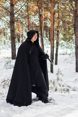 A warrior in black quilted clothes, in a black cloak with a hood on his head and a long sword in his hands against the background of winter forest and snow.