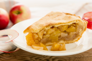 Homemade apple pie piece on  white plate with raw apples on wooden table background