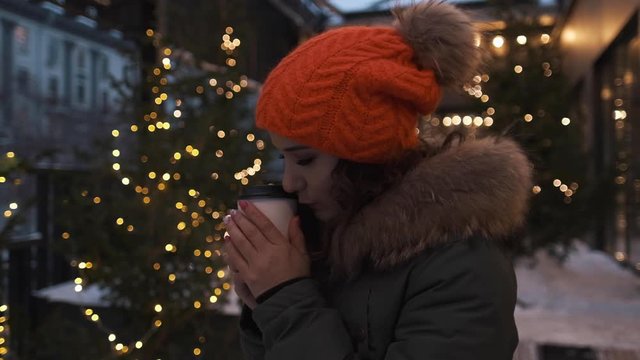 Portrait of an attractive girl in a bright hat with coffee in her hands in winter against the background of lights she warms her hands