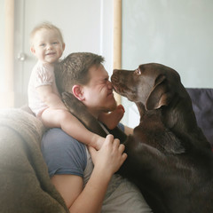Young caucasian father with cute smiling little baby girl sitting on his shoulders and pet labrador...