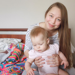 Young beautiful woman holding her sweet little baby, smiling, sitting on a bed