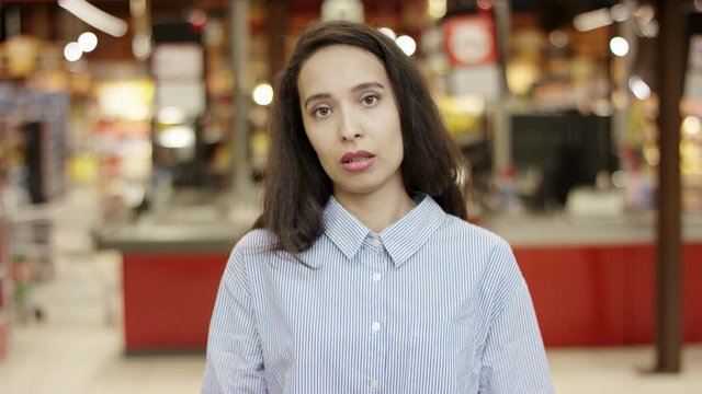 Lovely girl in supermarket being frustrated, feeling unhappy, rising hands up to show reproach and disappointment, shrugging shoulders in helpless gesture with people moving in blurred background.