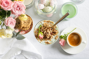 Breakfast concept. Sunday brunch. Light granola with Greek yogurt in a glass goblet close-up, coffee, light sweets and flowers on a snow-white tablecloth. Top View Image