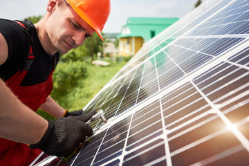 Young technician inspecting solar energy on a plot near the house. A man is wearing an orange uniform, hard hat, black t-shirt and gloves on a warm day. Alternative energy sustainable concept