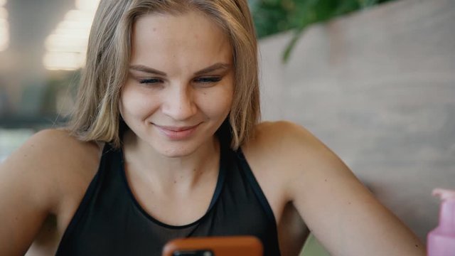 hipster young woman using smart phone to surf internet in gym cafe after workout. smiling female chats. concept of modern world, technology, waiting, business.