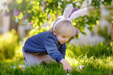 Fototapeta premium Pretty little boy wearing bunny ears hunting for easter eggs in spring park on Easter day.