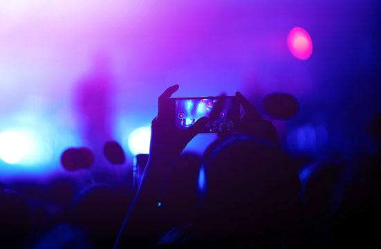 Hand with a phone records live music festival. People taking photographs with smart phone during a public music concert. Crowd raising their hands, dancing and enjoying great the concert.