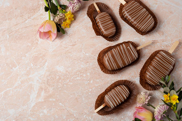 Chocolate pop cake in the form of popsicle with pink and yellow spring flowers on a marble background. Top View Image.