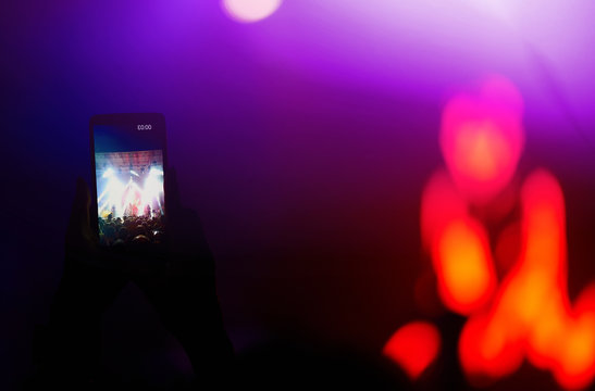 Hand with a phone records live music festival. People taking photographs with smart phone during a public music concert. Crowd raising their hands, dancing and enjoying great the concert.