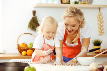 Little girl and her blonde mom in red aprons playing and laughing while kneading the dough in kitchen. Homemade pastry for bread, pizza or bake cookies. Family fun and cooking concept