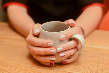 Closeup of womans hands holding a mug or cup of drink over light wooden table.