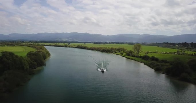 Drone shot of a river in New Zealand with boat and water ski rider. Nice green fields around and big mountains on the horizon.
