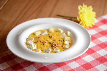 Creamy pumpkin soup topped with pumpkin seeds and cheese in a white bowl on a wooden table with plaid tablecloth.