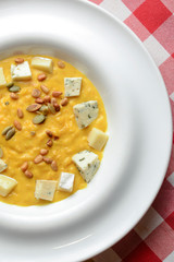 Creamy pumpkin soup topped with pumpkin seeds and cheese in a white bowl on a wooden table with plaid tablecloth.