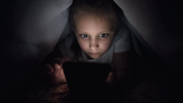 A young little girl is hiding under a blanket to use a digital tablet smartphone device late after sleep. Loneliness of young children and rescue on the phone.