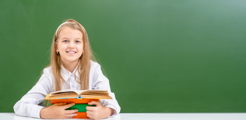 Smiling young girl sits with books near empty green chalkboard. Empty space for text