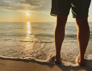 A man in shorts meets a beautiful sunset, sunrise. A warm sea wave with foam touches the legs. Sandy beach and a serene, tranquil place on the sea