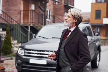 Fashionable senior man with gray hair and beard is outdoors on the street near his car
