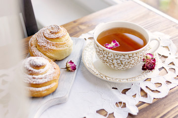 A cup of tea with roses buds, sweet buns with sugar powder on handmade white napkin and wooden table near window with day light. Beautiful breakfast or dessert time.