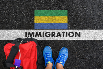 Man in shoes with bag standing next to line with word IMMIGRATION and flag of Gabon on asphalt road
