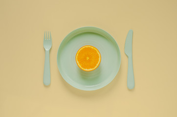 Cutting fresh orange fruit on green plate with knife and fork on yellow background. Minimal creative healthy diet food concept.