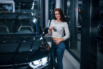 Young woman in casual clothes standing near modern black car with keys in hands