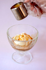 Chocolate syrup being poured over a scoop of vanilla ice cream in a bowl over pastel pink background.