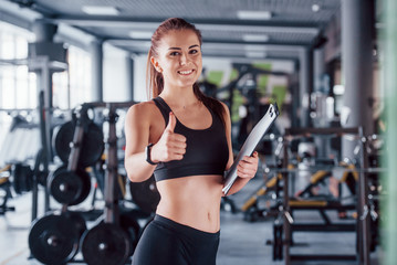 Fototapeta na wymiar Young female fitness personal trainer with notepad standing in the gym with thumb up