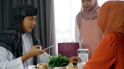 Young beautiful Asian Muslim man and women enjoying a relaxing moment in the coffee shop on a bright sunny day using mobile phone, smartwatch for mobile payment and QR code scan
