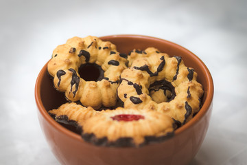 Crispy dough cookies stuffed with strawberry jam, with a chocolate-coated bottom. Inside a red pottery bowl. Against a blurry white background.