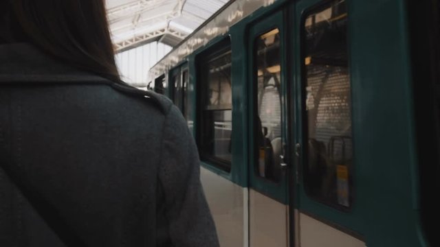 Rear view young woman walking along arriving metro train at Paris railway station platform, looking right slow motion.