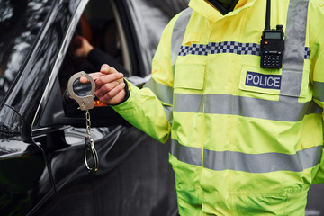 Male police officer in green uniformstanding with handcuffs near automobile